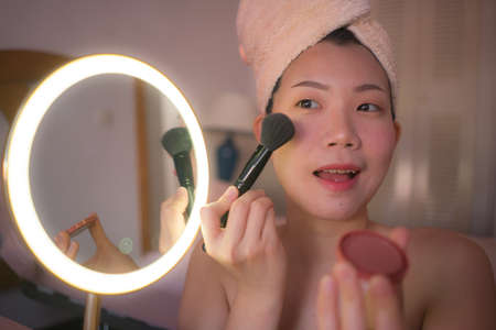 pretty Asian woman applying facial makeup - lifestyle portrait of young happy and beautiful Korean girl at home brushing her face applying beauty treatment in mirrorの写真素材