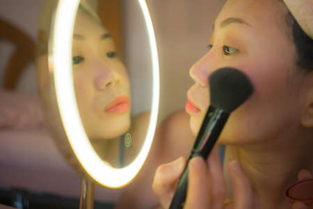 pretty Asian woman applying facial makeup - lifestyle portrait of young happy and beautiful Korean girl at home brushing her face applying beauty treatment in mirrorの写真素材