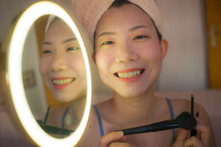 pretty Asian woman applying facial makeup - lifestyle portrait of young happy and beautiful Korean girl at home brushing her face applying beauty treatment in mirrorの写真素材