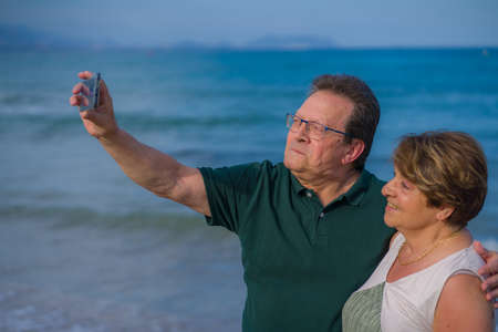 modern sweet and loving hispanic mature couple taking selfie portrait- senior retired husband and wife on their 70s enjoying beach walk relaxed and happy celebrating love togetherの写真素材