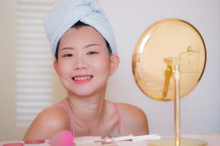 young beautiful and happy Asian Chinese woman with towel head wrap applying makeup looking to mirror smiling cheerful preparing for dating in beauty fashion conceptの写真素材