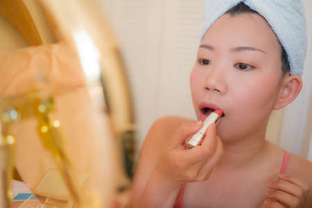 young beautiful and happy Asian Chinese woman with towel head wrap applying makeup looking to mirror smiling cheerful preparing for dating in beauty fashion conceptの写真素材