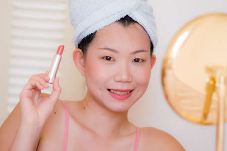 young beautiful and happy Asian Korean woman with towel head wrap applying makeup looking to mirror smiling cheerful preparing for dating in beauty fashion conceptの写真素材