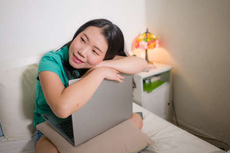 young beautiful Asian woman with laptop on bed -  lifestyle home portrait of young pretty and happy Korean student girl thinking and daydreaming with computer on bedroomの写真素材