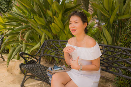 young beautiful and happy Asian woman on park bench - lifestyle portrait of Attractive Korean girl in Summer dress resting during tourist walk tourの写真素材