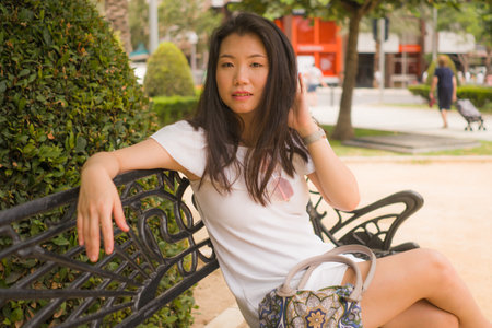 young beautiful and happy Asian woman on park bench - lifestyle portrait of Attractive Korean girl in Summer dress resting during tourist walk tourの写真素材