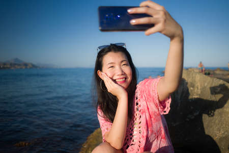 young happy and beautiful Asian woman taking selfie with mobile phone by the sea - Chinese girl enjoying summer holidays at the beach relaxed taking self portraitの写真素材
