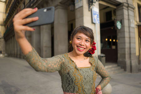 young happy and beautiful Asian woman wearing traditional Balinese kebaya dress taking selfie - Indonesian girl in Bali clothes walking on city street during holidays in Europeの写真素材