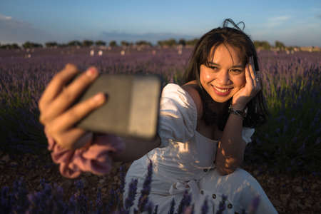 outdoors romantic portrait of young happy and attractive woman in white summer dress taking selfie with mobile phone at beautiful lavender flowers field in holiday conceptの写真素材