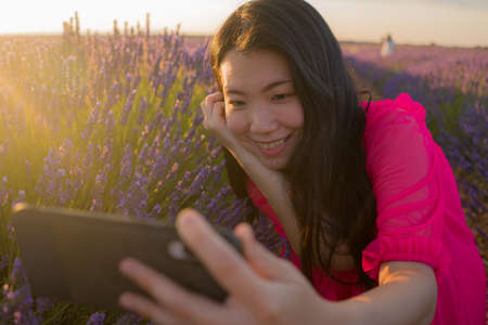 young happy and beautiful Asian Japanese woman in Summer dress taking sunset selfie with mobile phone at purple lavender flowers field in nature and romantic beauty conceptの写真素材