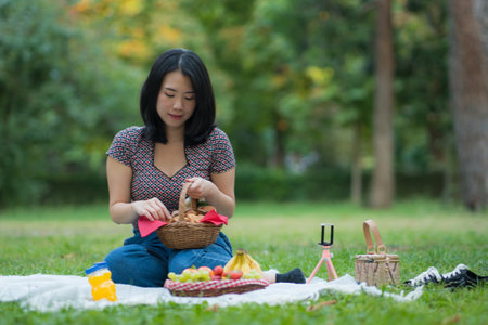 Autumn picnic at city park - lifestyle portrait of young happy and beautiful Asian Chinese woman sitting grass with blanket and fruit basket enjoying relaxed a weekend picnicの写真素材