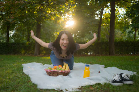 Autumn picnic at city park - lifestyle portrait of young happy and beautiful Asian Chinese woman sitting grass with blanket and fruit basket enjoying relaxed a weekend picnicの写真素材