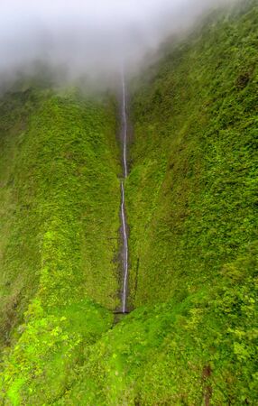 Spectacular aerial view of high waterfall in the green mountains shrouded in fog under a cloudy sky at Na Pali Coast, shot taken from a helicopter, Kauai, Hawaiiの写真素材
