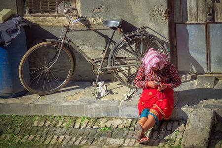 Bhaktapur, Nepal - 11/21/2017: Everyday life, woman sitting on the curb clipping her nails, an old bike leaning against the wall of her home.のeditorial素材