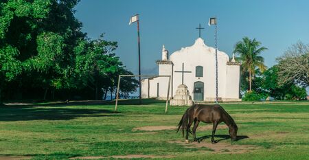 Horse is eating grass in front of Quadrado Church, there is a cross, a goal post and to tall flags...