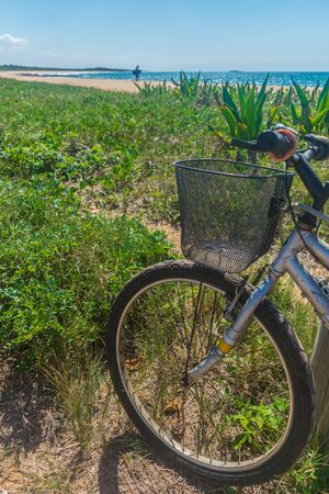 Front part of a bicycle standing in the beach, the bike has a basked fixed in front of it, a meadow of grass covers the half of the shore, the blue ocean is seen in the horizon while a man walksの写真素材