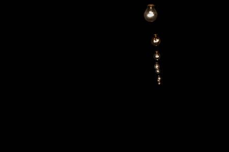 Dark and profound overview of a sequence of aligned incandescent light bulbs forming a line and hanging into the air going inwards into the shadows at the right corner of the pictureの写真素材