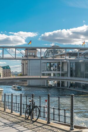 Scenic view of a bicycle leaning in a metal fence, with a boat riding in the Spree river and the metal architecture of the german parliament with national flags on its roof in the backgroundの写真素材