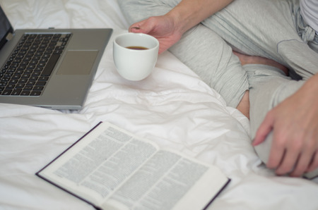 Woman with crossed legs, reading book and drinking coffee, computer in bed. Seen from the top laterally.の写真素材