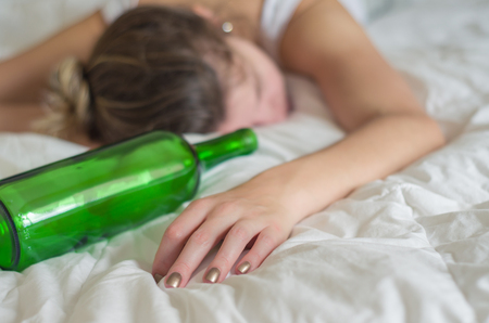 Great concept of alcohol abuse. Young woman, blond hair, fainted in bed after drinking too much alcohol. Glass of wine in hand, bottle of wine.の写真素材