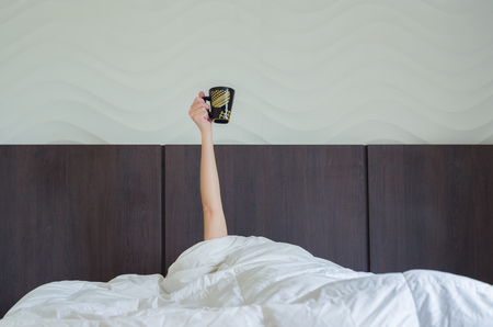 Drinking coffee early tomorrow. Woman in bed, under duvet, with raised arm holding coffee cup.の写真素材