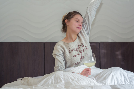 Young blonde woman stretching on bed with glass of wine in hand, waking up happy.