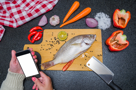 Young woman consulting recipe for fish on smartphone, fish and ingredients on the table. Preparing a...
