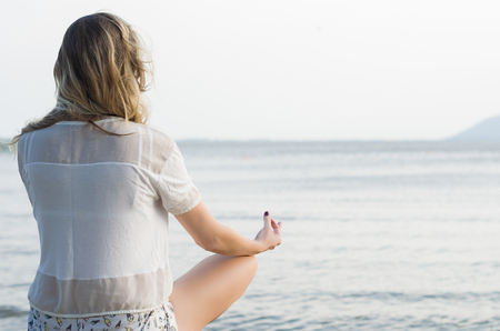 Young woman practices yoga and meditates in the lotus position on the beach, facing the water. Great concept of meditation and relaxation.の写真素材