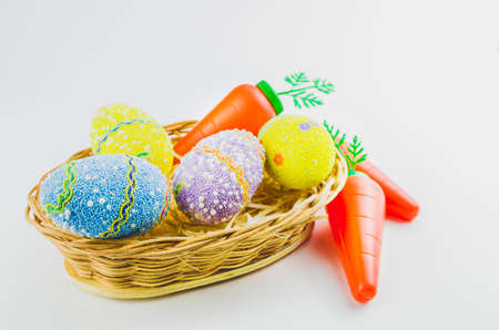 Easter colored eggs and carrots in a basket isolated on a trendy white background. Minimal easter concept. Easter card with copy space for text.の写真素材