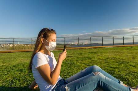 Young woman sitting on the grass with a protective mask during the SARS CoV-2 pandemic season ,...