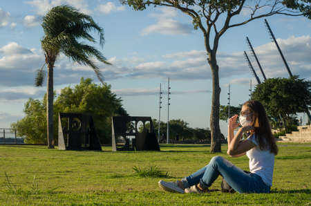 Young woman sitting on the grass with a protective mask during the SARS CoV-2 pandemic season  on...