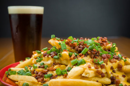 Tasty close-up of french fries, cheddar and bacon, pint of beer on wooden and black background