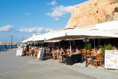 Chania, Crete - Greece - October 20 2021 :  Charming pavement taverna at the historic Venetian harbor  Holiday makers enjoy the summer sun  Landscape aspect shot with copy spaceのeditorial素材