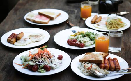 Breakfast set is on the table with sausage, vegetable salad, omelette, ham, bread, fruit, milk,...
