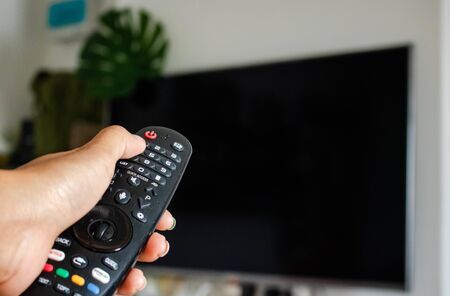Watching Television Concept. Hand using a remote control in front of the TV screen. Relaxation in Modern Living Room. Focus on Remote. copy space.の写真素材