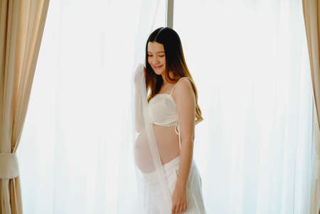 Young beautiful pregnant woman standing of happy pregnant woman with big belly at window. Smiling pregnant woman.の写真素材