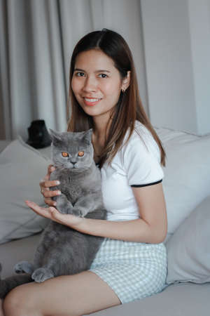 Happy beautiful young brunette woman holding and hugging her cute British shorthair cat on couch.の写真素材