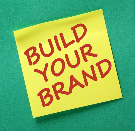 The words Build our Brand in red text on a yellow sticky note posted on a green notice board as a reminderの写真素材