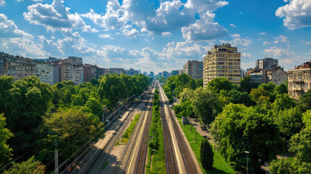 Vibrant urban landscape featuring railway tracks flanked by lush greenery under a bright blue sky with fluffy clouds. A captivating blend of nature and city life.の素材