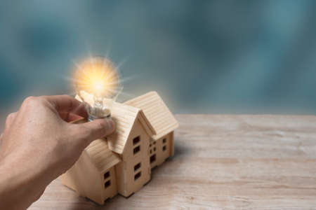 A man's hand holding a light bulb and a wooden house model from a model on a wooden table. Real estate concept, copy space for various textsの写真素材