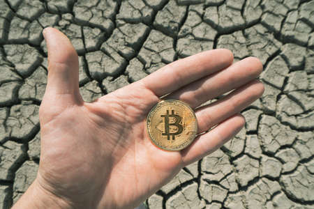 BITCOIN OVER DRY LAND IN HAND SYMBOLIZING GLOBAL WARMING AND CRYPTO MININGの写真素材