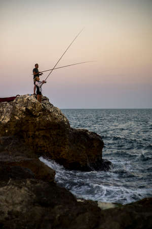 MAN AND WOMAN ROCK FISHING ON THE SEA COAST DURING SUNSET.の写真素材