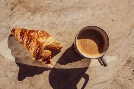 This scene features a croissant half eaten and an untouched coffee on a marble table in the morning with both shadows.の写真素材
