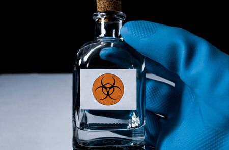Black background, cork lead small bottle with white label and orange toxic international symbol held by a right hand with a blue glove.の写真素材