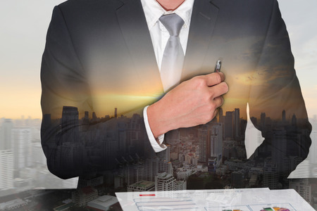Double exposure of businessman holding a pen on the pocket, cityscape and sunset as business and analysis concept.の写真素材