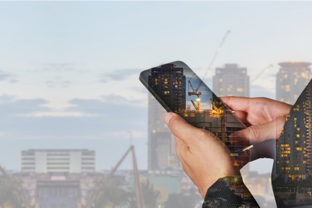 double exposure of woman hand hold and touch screen smart phone, cellphone, construction crane and building in the evening, twilight as business, industrial, communication and technology concept.の写真素材