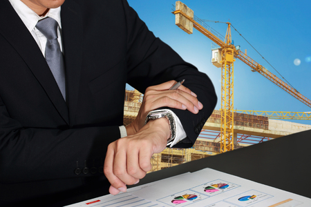 Businessman look a watch in appointment on blur crane construction, urban and city isolated background as business and take a time concept.の写真素材