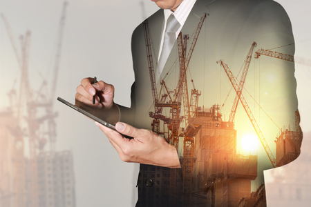 Double exposure of businessman hand hold tablet, construction crane, building and sunset in the evening as business, technology, communication and industry concept.の写真素材