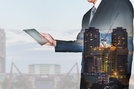 Double exposure of businessman looking the tablet, construction crane and building in the evening, twilight as business, technology, communication and industrial concept.の写真素材