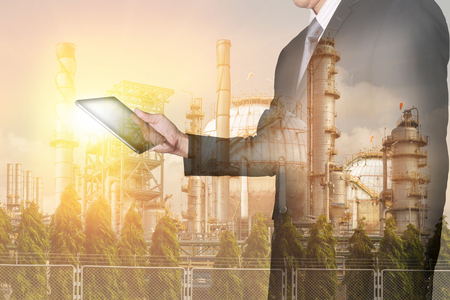 Double exposure of businessman looking the tablet, oil fuel, electric generating factory or plant with sunset as business, technology, communication, industry and energy concept.の写真素材
